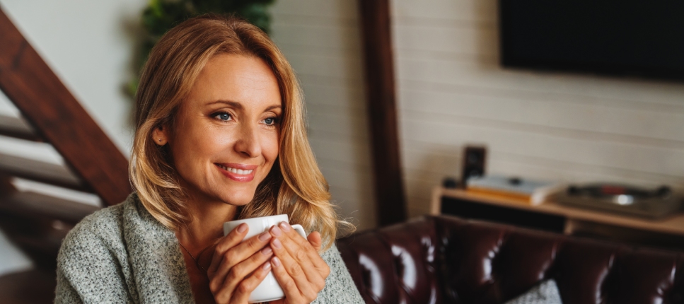 A smiling woman with blonde hair sits in a cozy living room, holding a white mug with both hands close to her chest