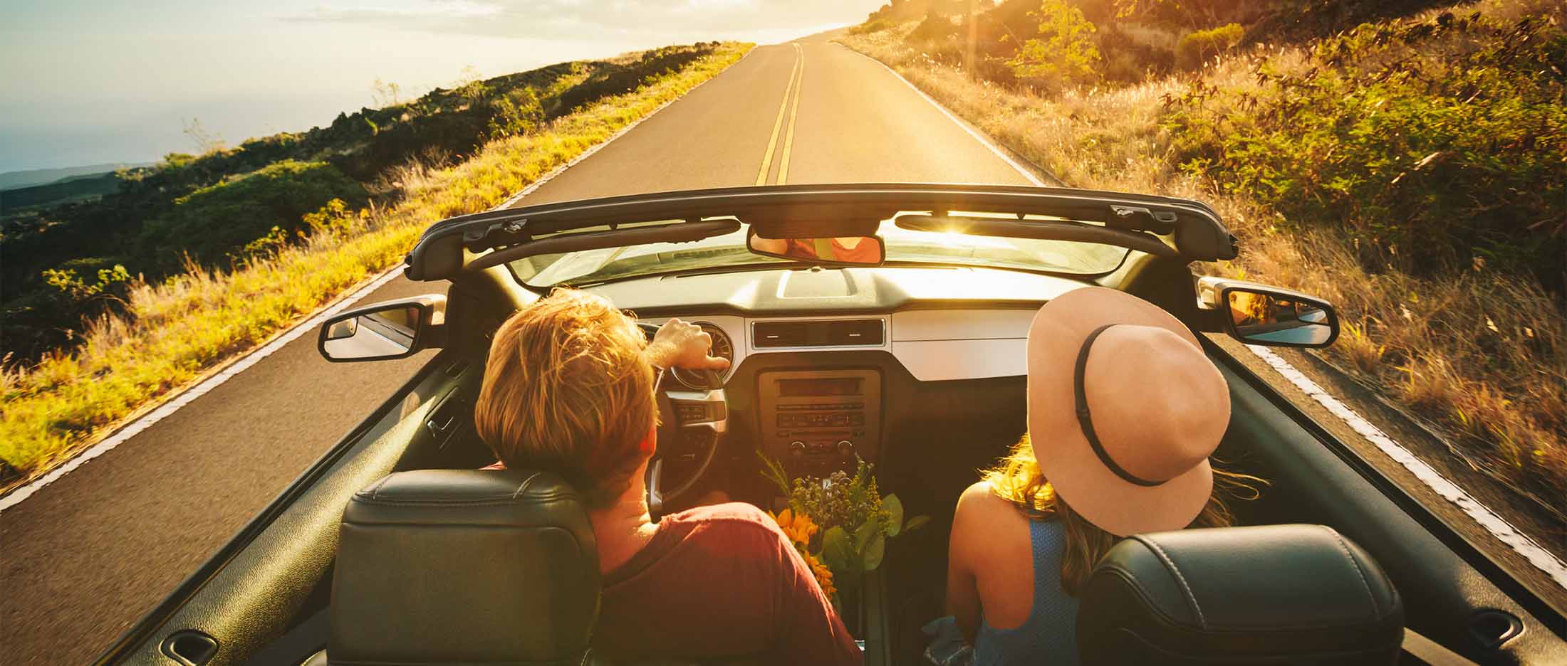 A couple in a convertible drives down a winding road toward a golden sunset, viewed from the back seat
